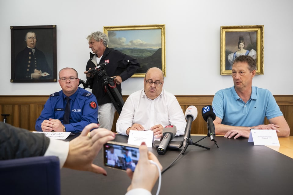 Drei Männer sitzen an Bürotisch; vor ihnen sind Pressemikrofone aufgereiht