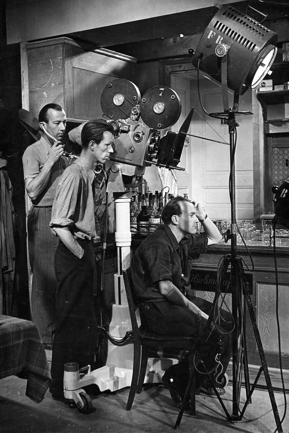 Drei Männer (zwei stehend, einer sitzend auf einem Stuhl) mit der Kamera in der Mitte und einem Filmscheinwerfer neben ihnen.