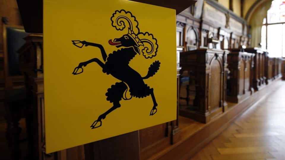 Kantonsratssaal mit holzverkleideten Pulten. Vorne im Bild ein gelbes Wappen mit Schafsbock, dessen Hoden zu sehen sind