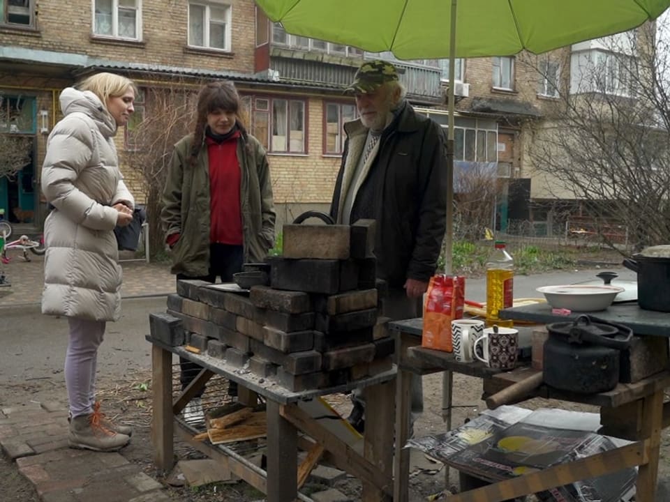 Luzia Tschirky steht auf der Strasse neben einem älteren Mann und einer jüngeren Frau, die am offenen Feuer etwas kochen