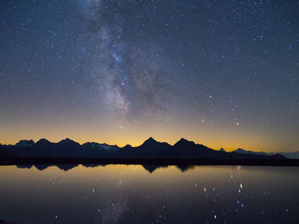 Ein spiegelglatter Bergsee reflektiert den klaren Sternenhimmel.