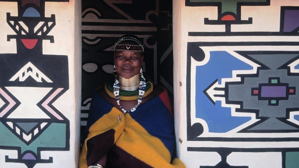 Schwarze Ndebele-Frau steht in einem Hauseingang. Es ist mit geometrischen Mustern gestaltet, wie ihre Kleidung.