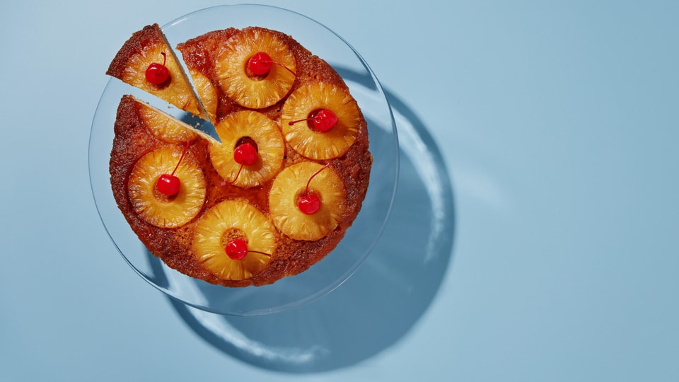 Mit Ananasscheiben belegter Kuchen auf einer Glasplatte vor hellblauem Hintergrund