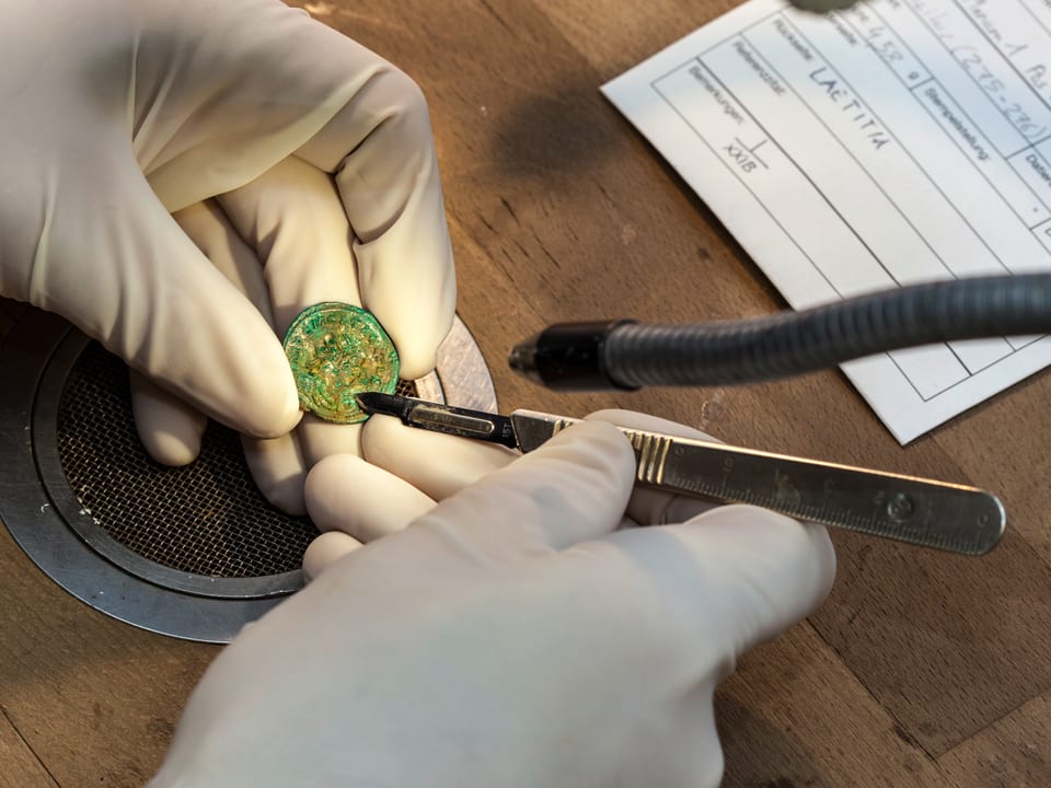 Zwei in Handschuhe gepackte Hände, ein Griffel, mittendrin eine alte Münze