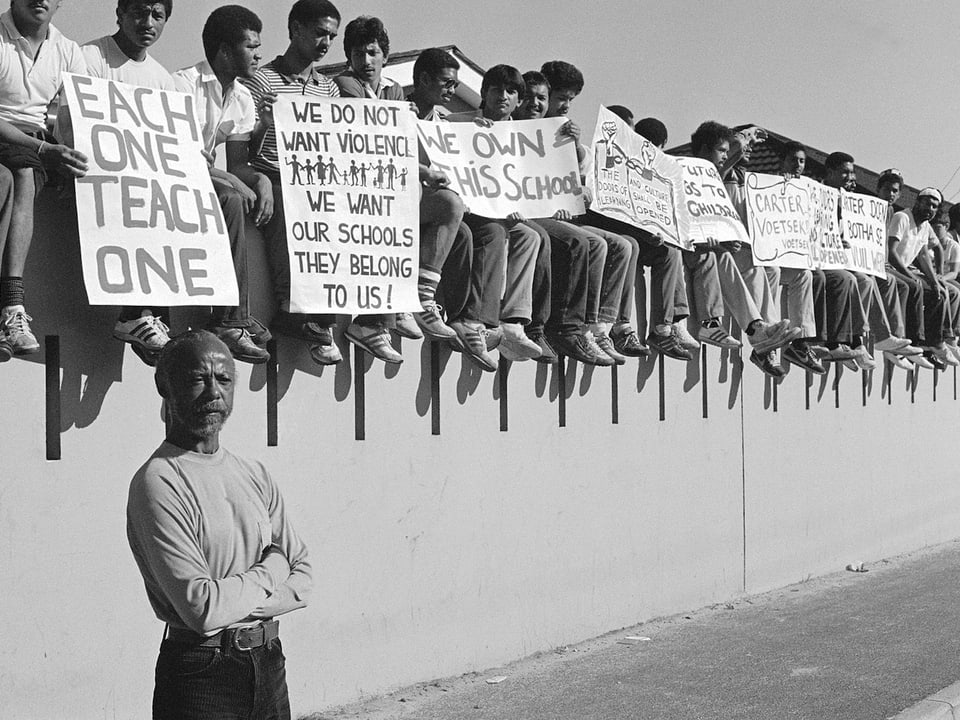 James Matthews protestiert zusammen mit Schülern. Sie sitzen alle auf einer Mauer und halten Plakate.