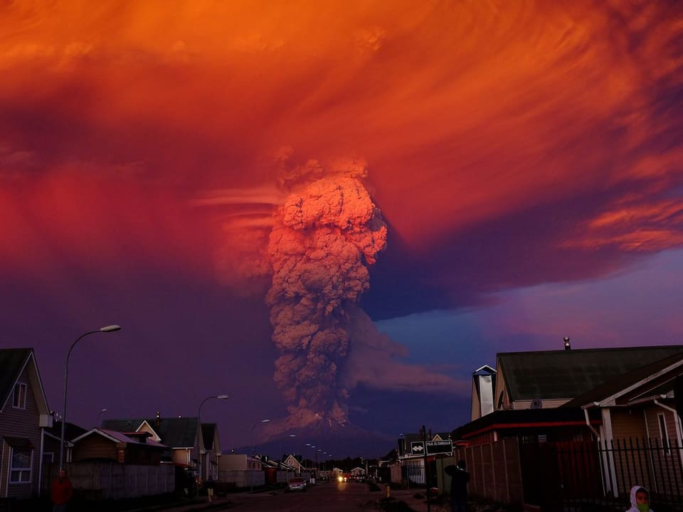 Eine rote Aschewolke über einem Vulkan mit einer Häuserzeile im Vordergrund.