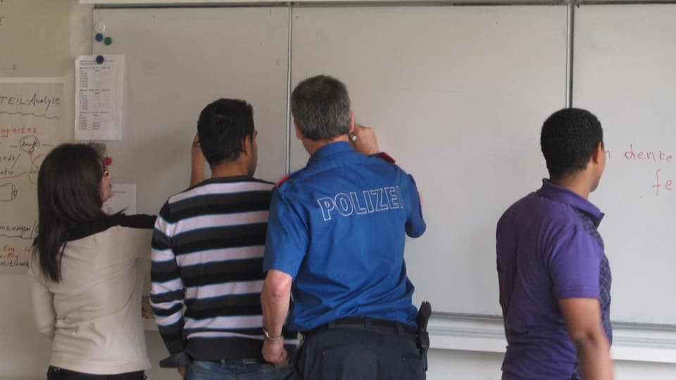 Ein Whiteboard, davor ein Mann in Polizeiuniform und drei Personen, die alle an die Tafel schreiben.