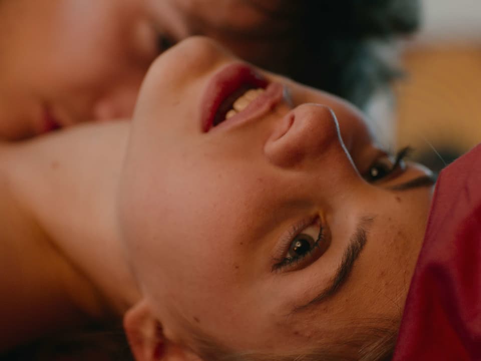 Filmszene: Gesicht einer jungen Frau, die auf einem Bett liegt. EIn Mann küsst ihren Hals.