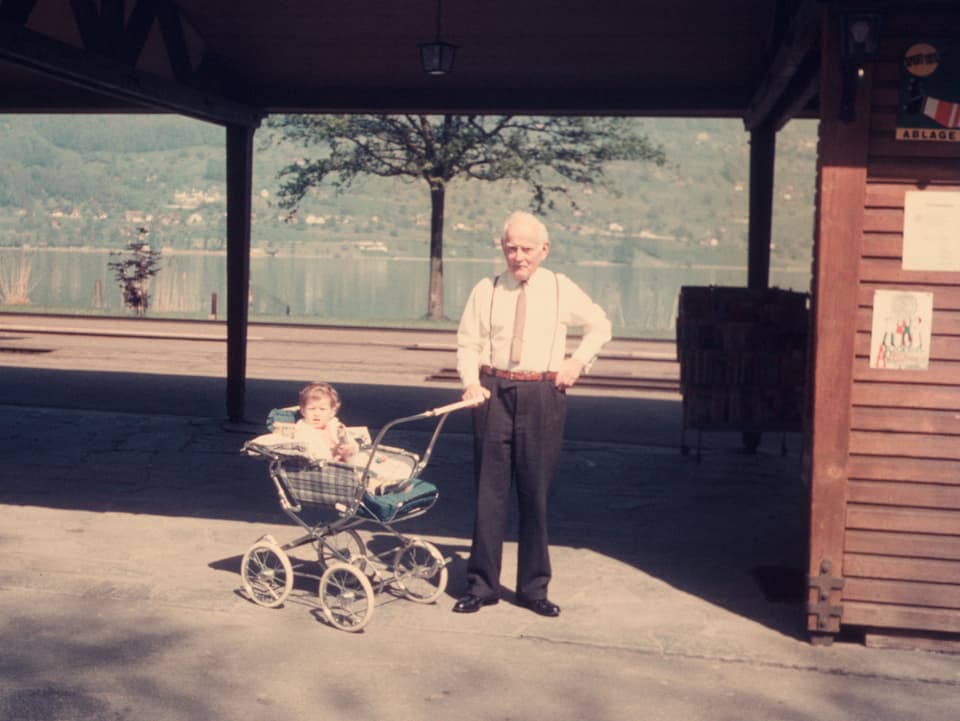 Ein älterer Mann hält einen Kinderwagen, darin sitzt ein kleines Kind. 