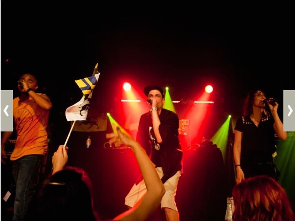 Zu sehen ist ein Rapper auf einer Bühne, assistiert von zwei Backgroundsängern, im Vordergrund schwingt einer der Fans eine kleine Bündner Fahne.