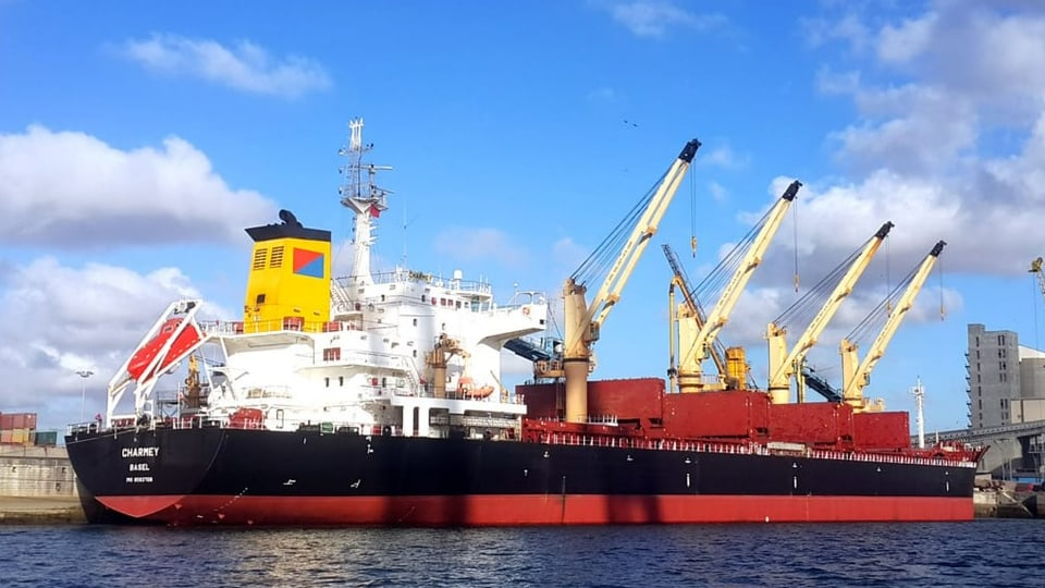 Ein Schiff im Hafen. Name und Standort «Basel» ist am Heck zu lesen. Vier Ladekrane ragen im Hintergrund.