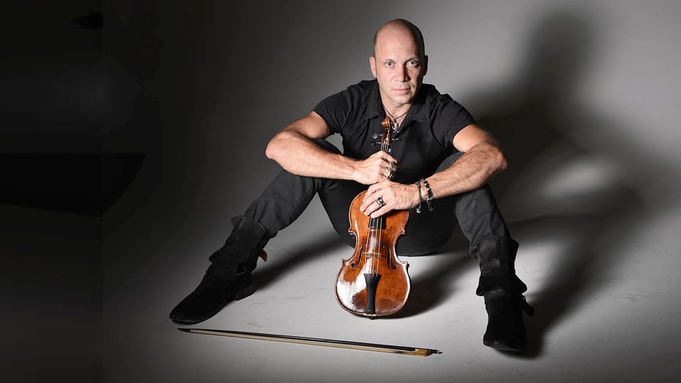 Der schwarz gekleidete Stefano Montanari sitzt am Boden mit gespreizten Beinen, in den Händen hält er eine Geige.