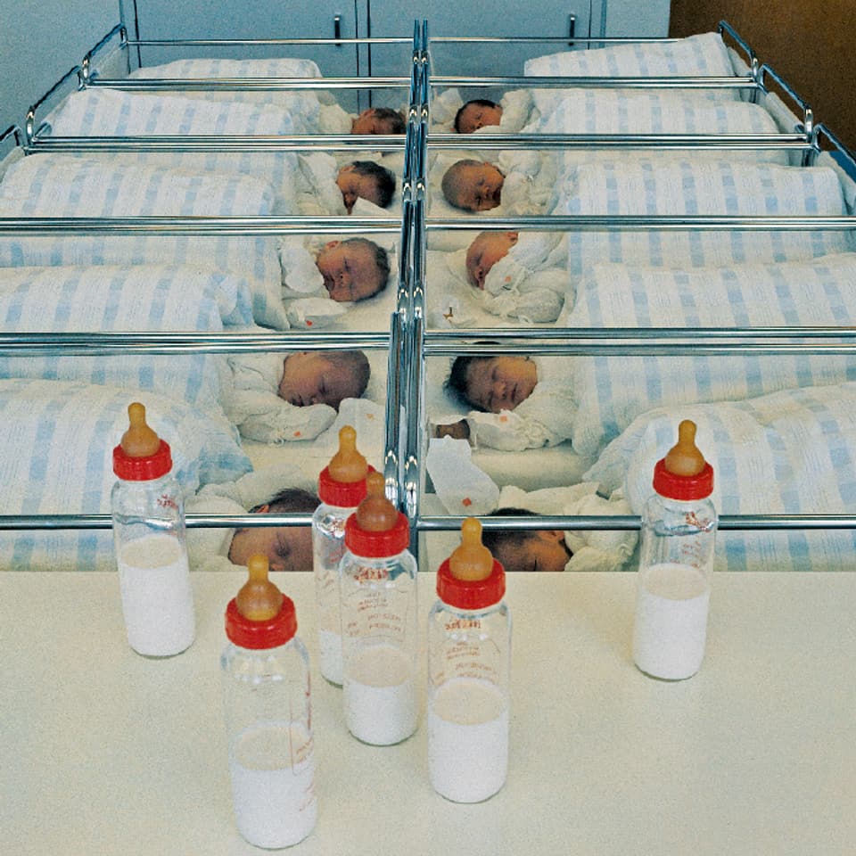 In einem Säuglingszimmer stehen zehn kleine Betten mit Säuglingen, davor ein paar Flaschen Milch.