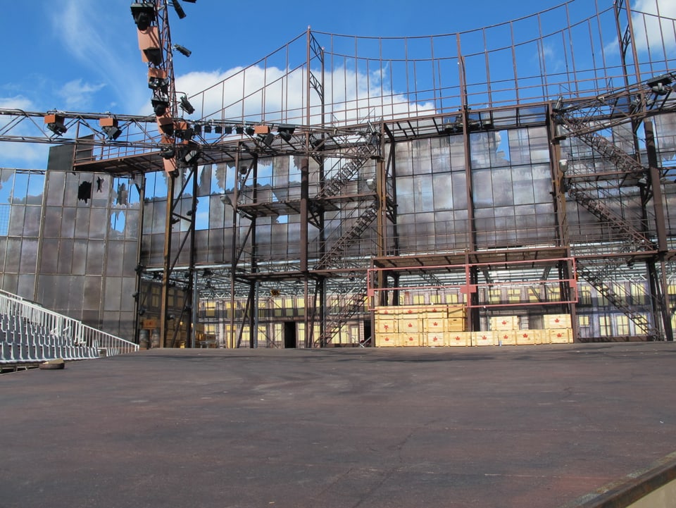 Blick auf die Bühne mit einer Eisenkonstruktion im Hintergrund, welche die Brücke darstellen soll. Darunter stehen helle Holzkisten als Kulisse. 