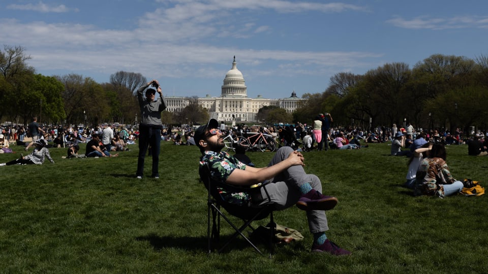 Viele Menschen sitzen im Gras vor dem Capitol in Washington DC und schauen in den Himmel. 