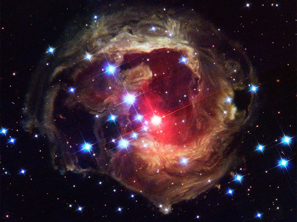 Eine Aufnahme des Hubble Teleskops. Es zeigt einen rötlichen Farbschleier aus abertausenden Punkten. Dazwischen scheinen Sterne, also kleine Sonnen ins Weltall.