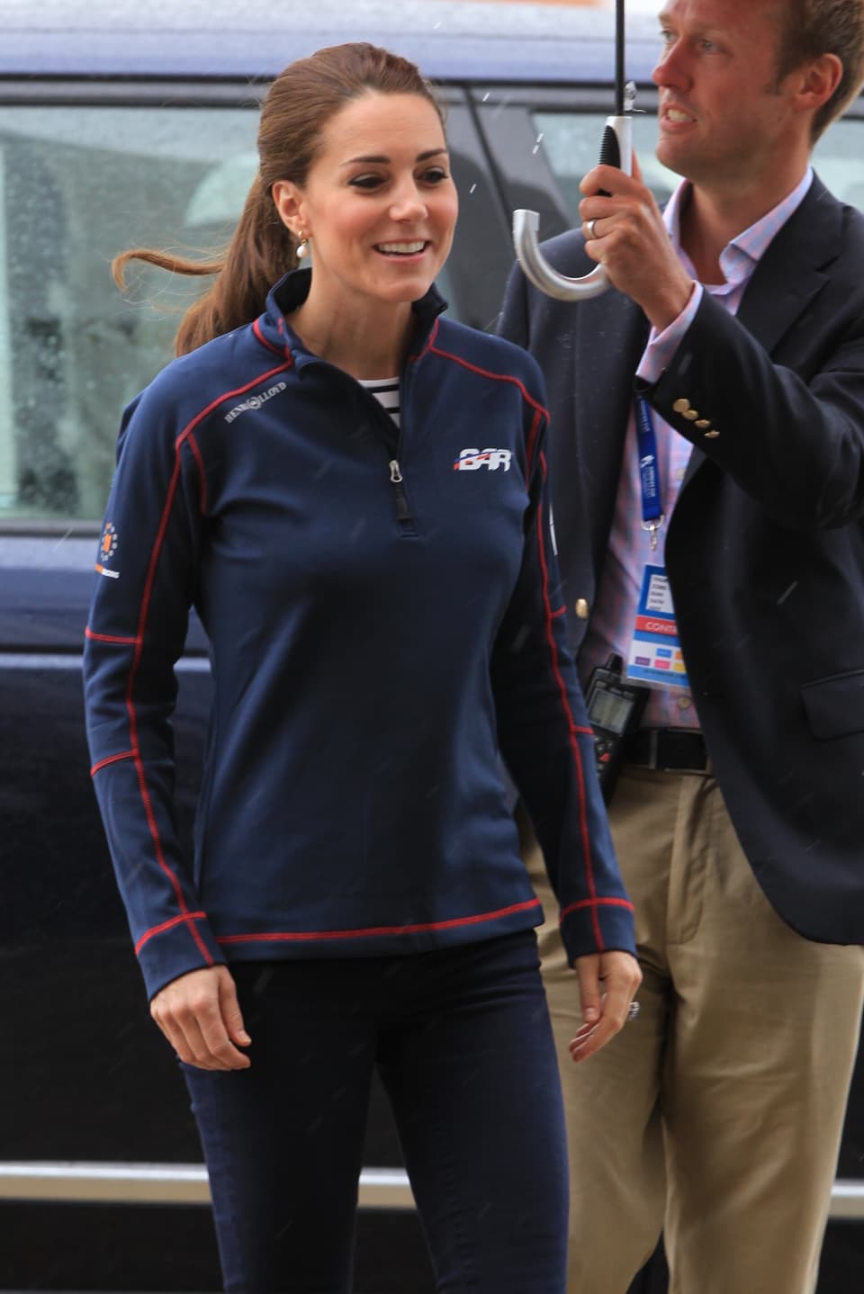 Herzogin Kate lächelt in blauer Fleece-Jacke, während ein Bediensteter für sie den Schirm hält. 