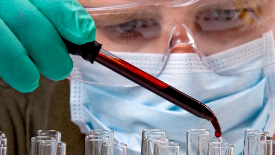 Ein Rechtsmediziner mit Schutzbrille und Mundschutz tröpfelt mit einer Pinzette Blut in kleine Glasgefässe, um eine Probe zu analysieren.