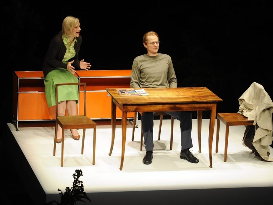 Ein Mann und eine Frau auf einer Bühne, er sitzt an einem Tisch, sie lehnt gegen ein Möbel