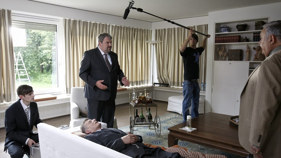 Four men stand around a man lying on the sofa. One of the men holds a telescopic microphone in his hand.