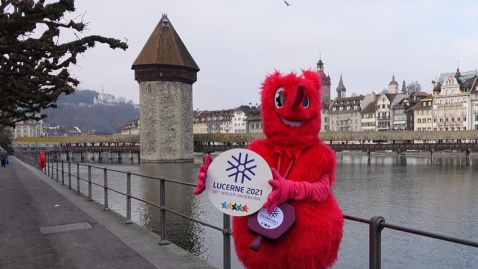 Ein rotes, haariges Maskottchen hält das Logo der Luzerner Winteruniversiade 2021, im Hintergrund die Kapellenbrücke.