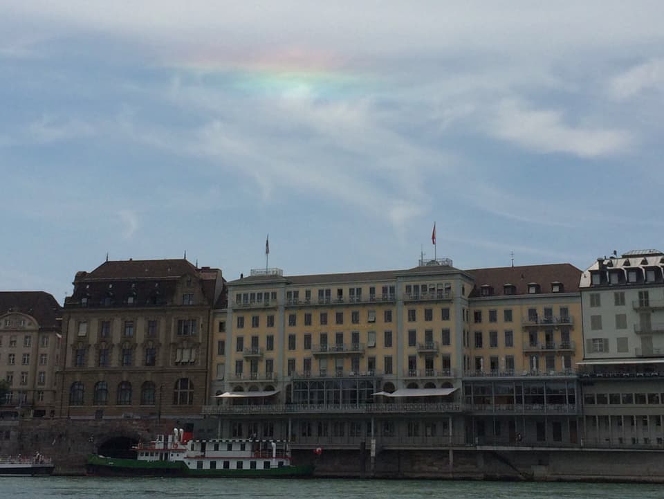 Im Vordergrund das Hotel Drei König in Basel mit dem Rhein. Darüber blauer Himmel mit Schleierwolken. Darin leuchtet ein Streifen in Regenbogen Farben.