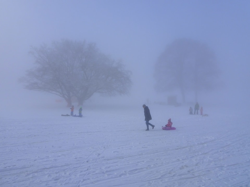 Kinder fahren bei Nebel über einen leicht geneigten Hang. Es liegt überall Schnee, die Bäume sind im Nebelgrau gerade noch zu erkennen.