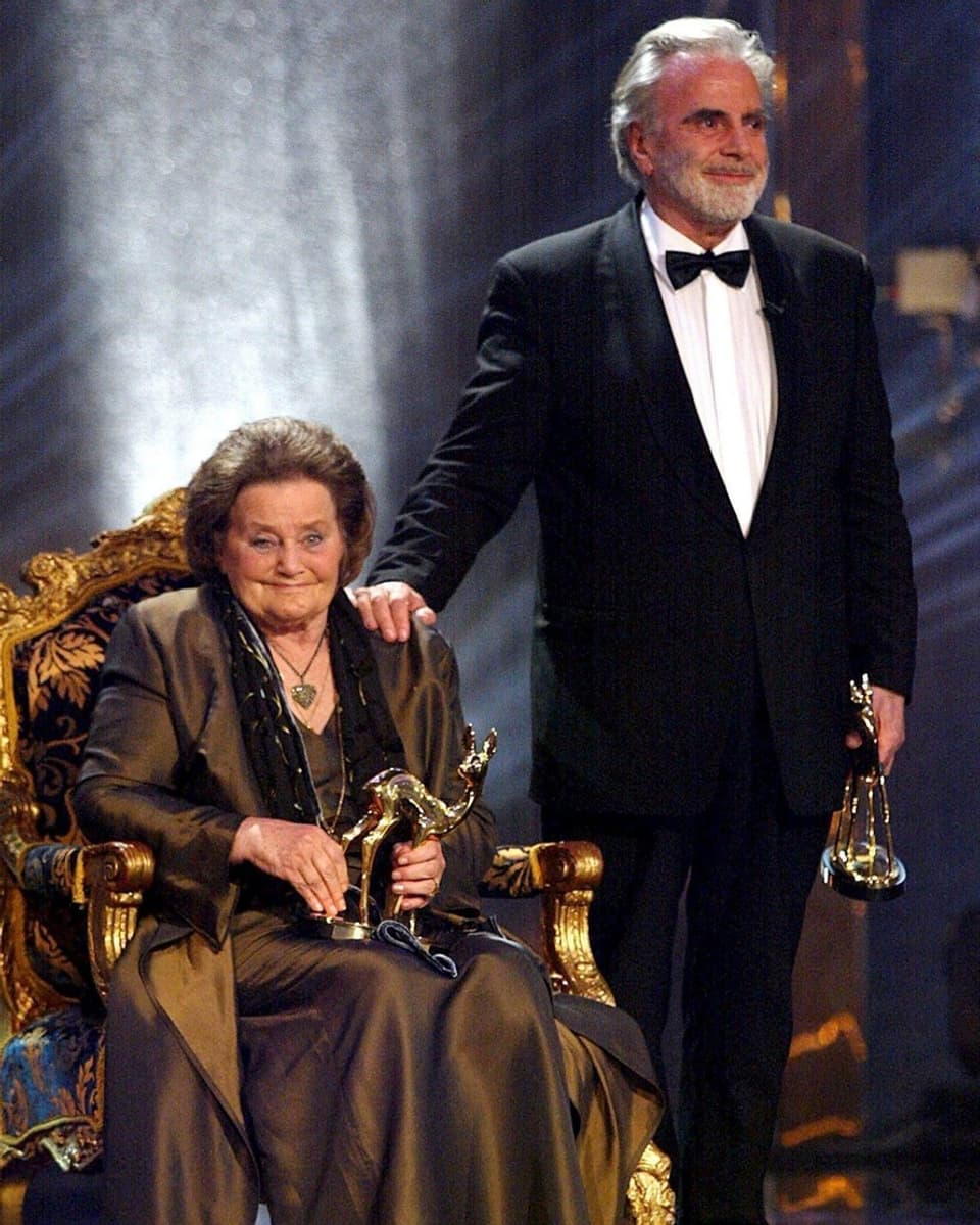 Maria Schell sitzend und Maximilian Schell stehend in Abendgarderobe mit einem Bambi-Award in der Hand