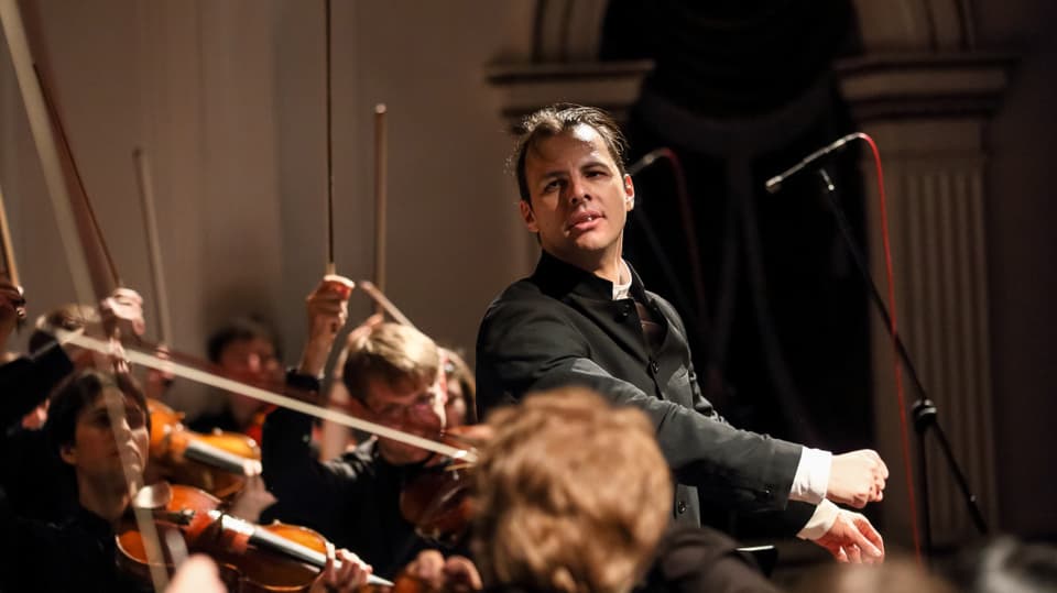 Junger Mann mit nach hinten gelegtem Haar steht am Dirigentenpult inmitten eines Orchesters.