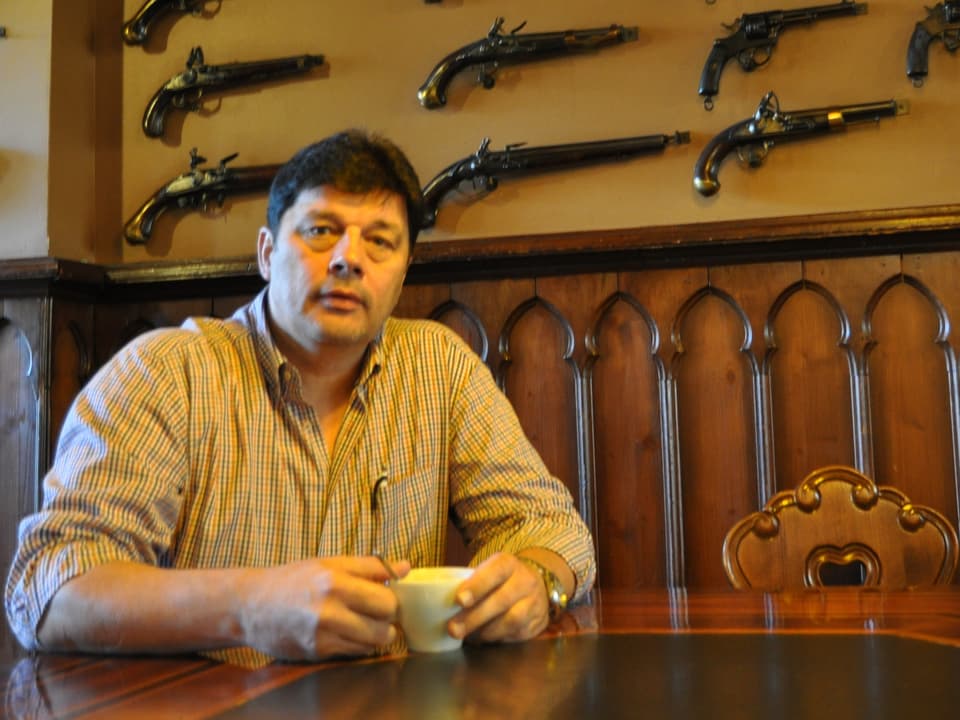 Mann mit Kaffeetasse an Tisch. Gewehre an der Wand.