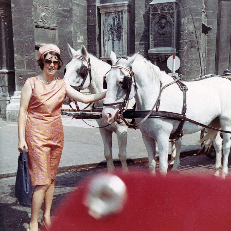 Die junge Frau trägt ein oranges Kleid mit passendem Hut und steht vor einem Zweispänner mit weissen Pferden.