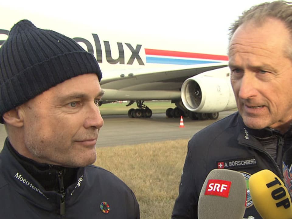Zwei Männer vor einem Transportflugzeug. Den beiden werden Mikrofone hingehalten.