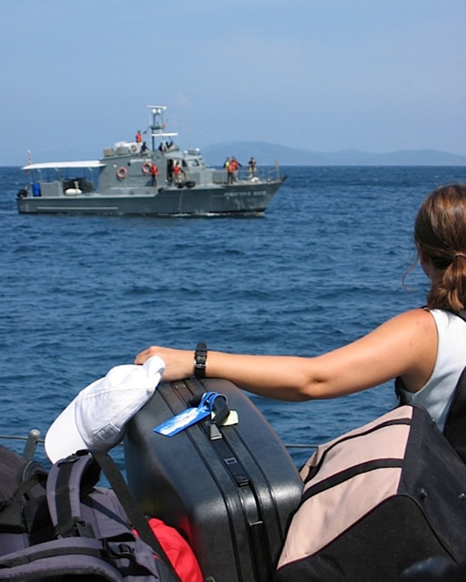Ein Armeeschiff auf dem Meer, im Vordergrund eine Frau mit Koffern.