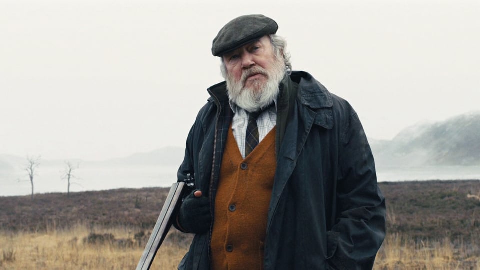 Ein alter Mann mit Bart und einem Gewehr im Arm steht auf einem nebligen Feld.