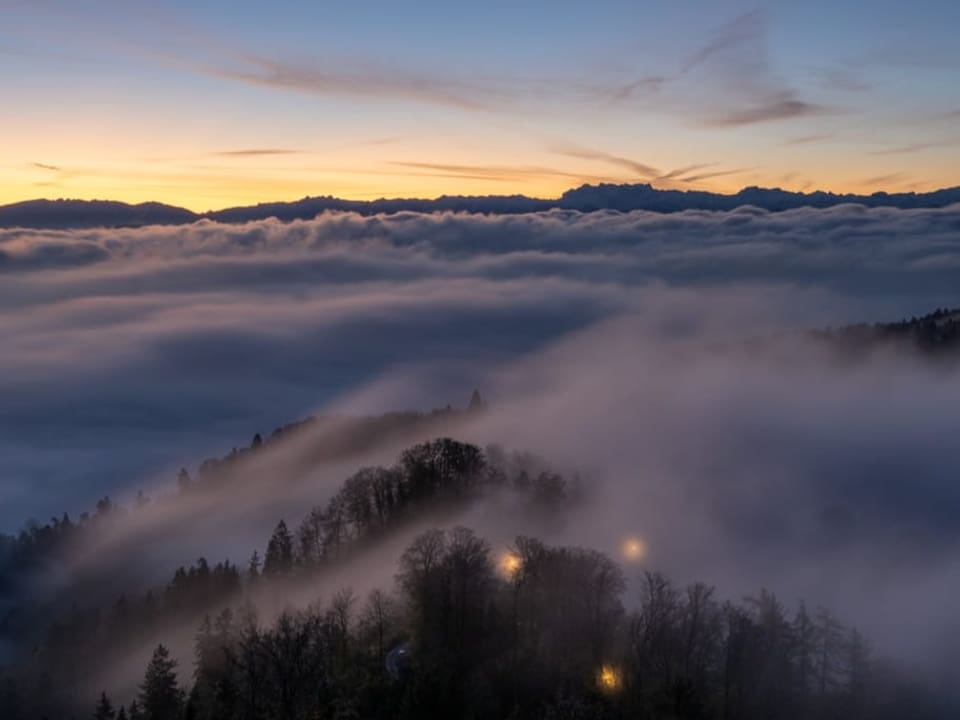 Nebelmeer mit Hügel, der aus dem Nebel ragt in der Dämmerung. Darüber hellblauer, klarer Himmel. 