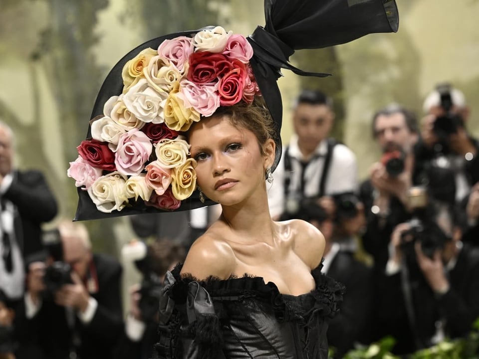 Model mit Blumenhut und schwarzer Kleidung bei einer Modeschau mit Fotografen im Hintergrund.