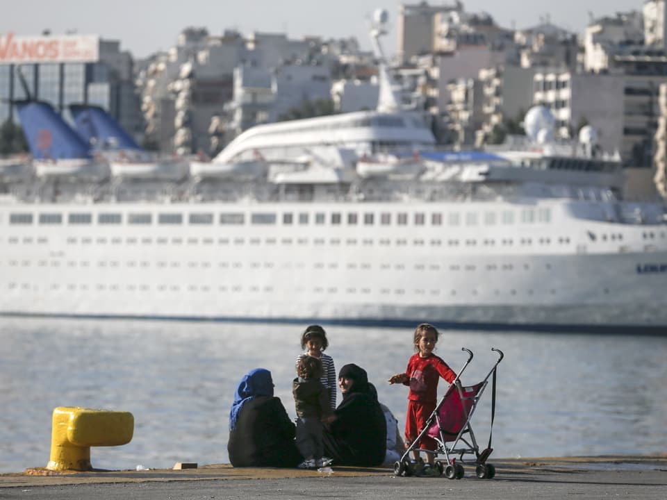 Eine Gruppe Frauen und Kinder sitzt am Pier, im Hintergrund eine Passagierfähre.