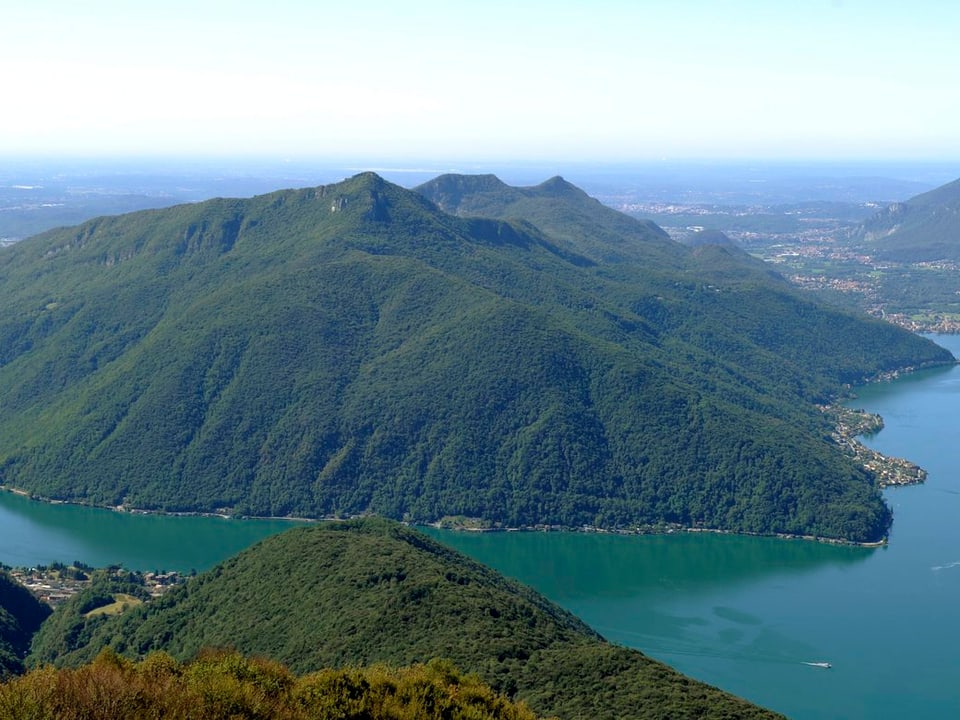 Grün bewachsener Monte San Giorgio mit Arm des Luganersees im Vordergrund.
