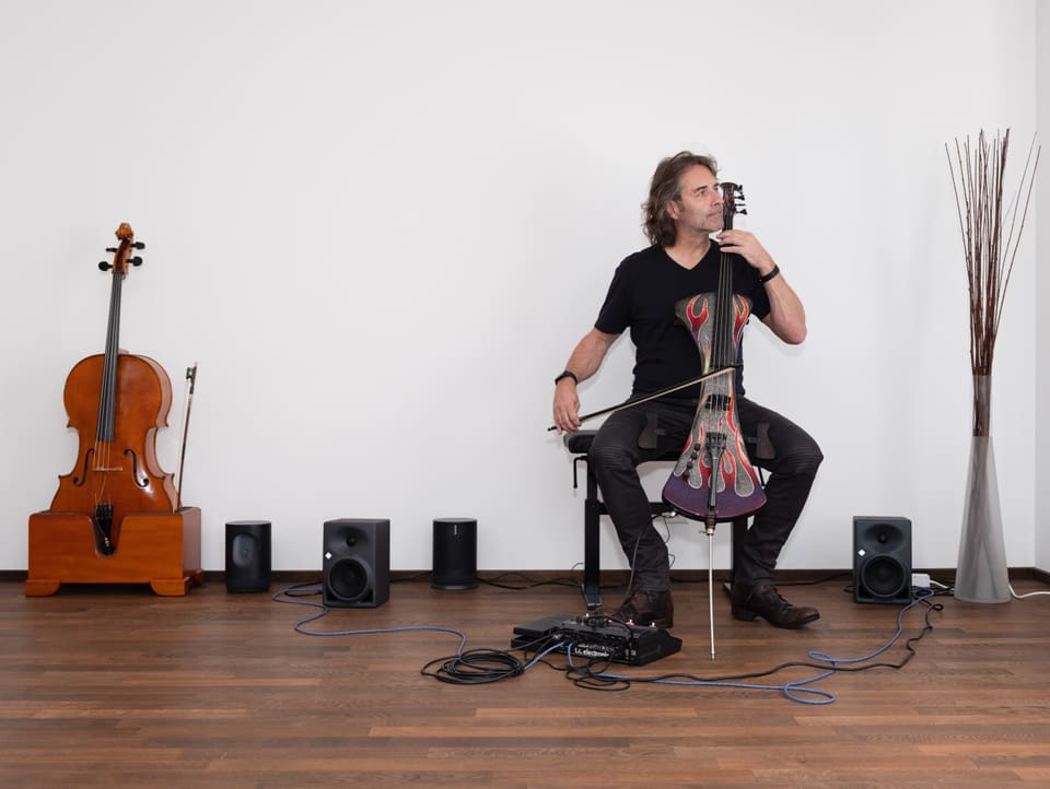 Ein Mann spielt im Sitzen an die Wand gelehnt ein Elektro-Cello, links neben ihm steht ein weiteres Cello an der Wand