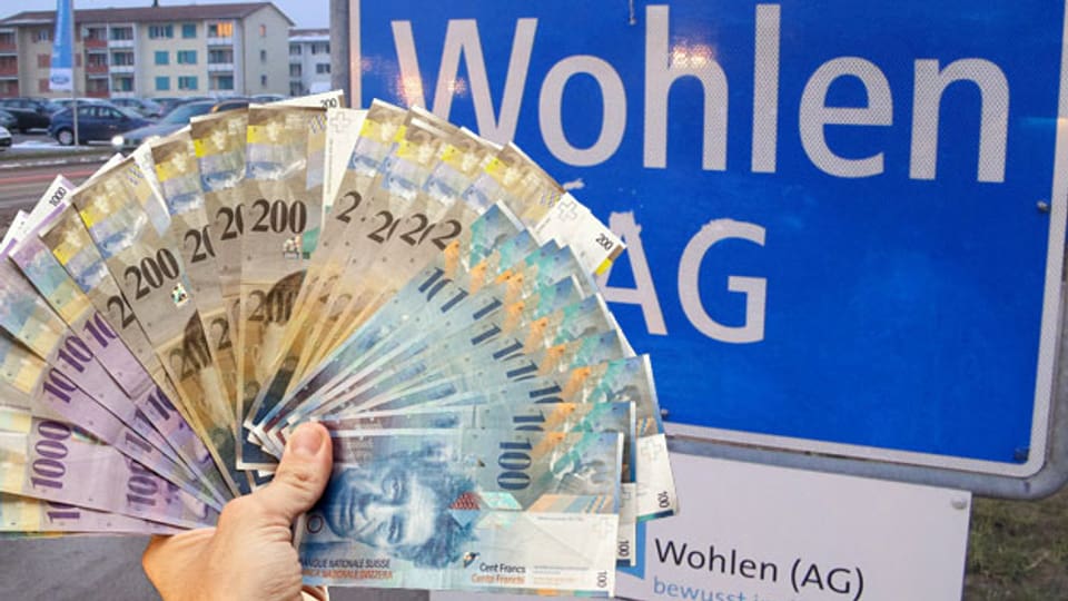 Eine Hand mit Banknoten vor dem Ortseingangsschild von Wohlen.