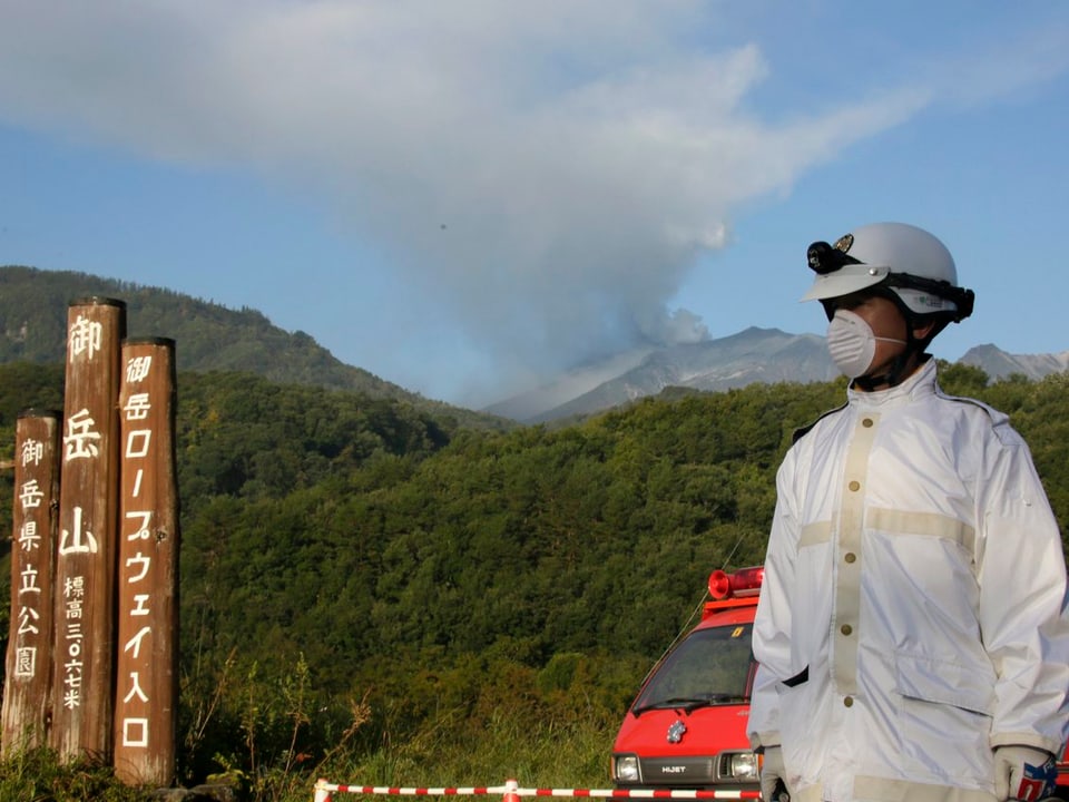 Ein Polizist mit Schutzanzug und Atemschutz sperrt die Zugangsstrasse zum Vulkan, der im Hintergrund raucht