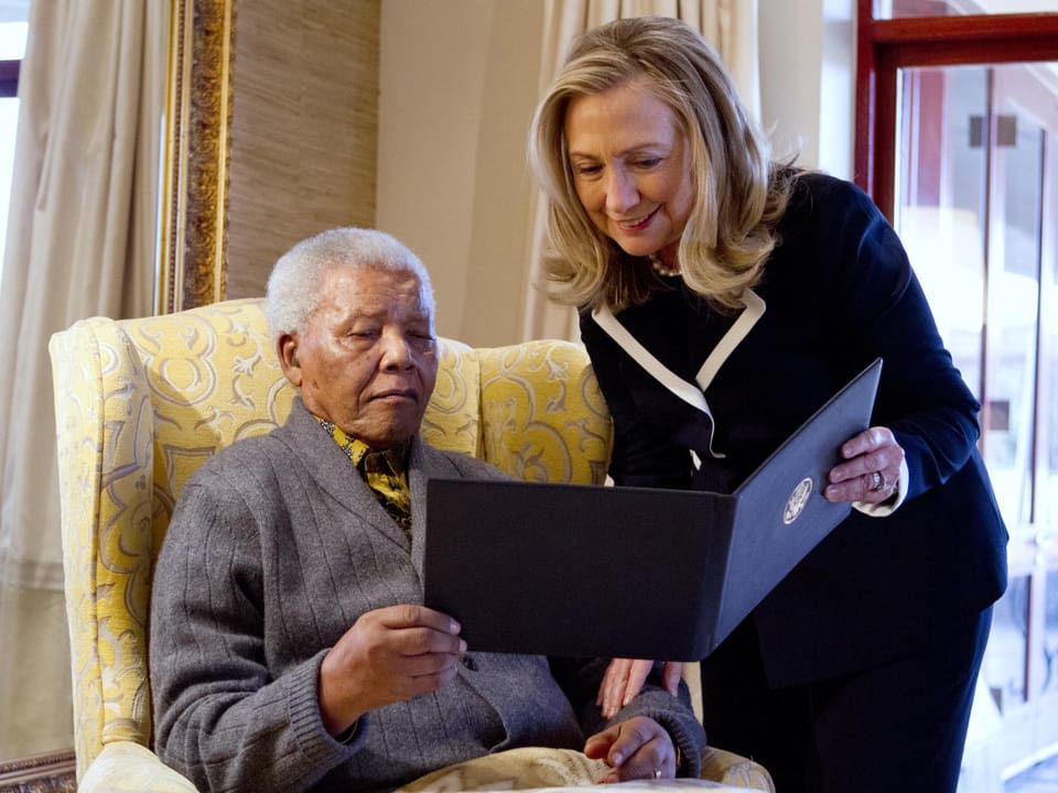 Hillary Clinton (rechts) neben Mandela. Sie sehen sich ein Buch an.