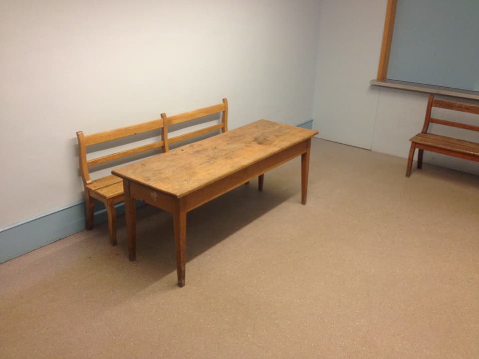 Holztisch mit Holzbank in ansonsten leerem Raum. 