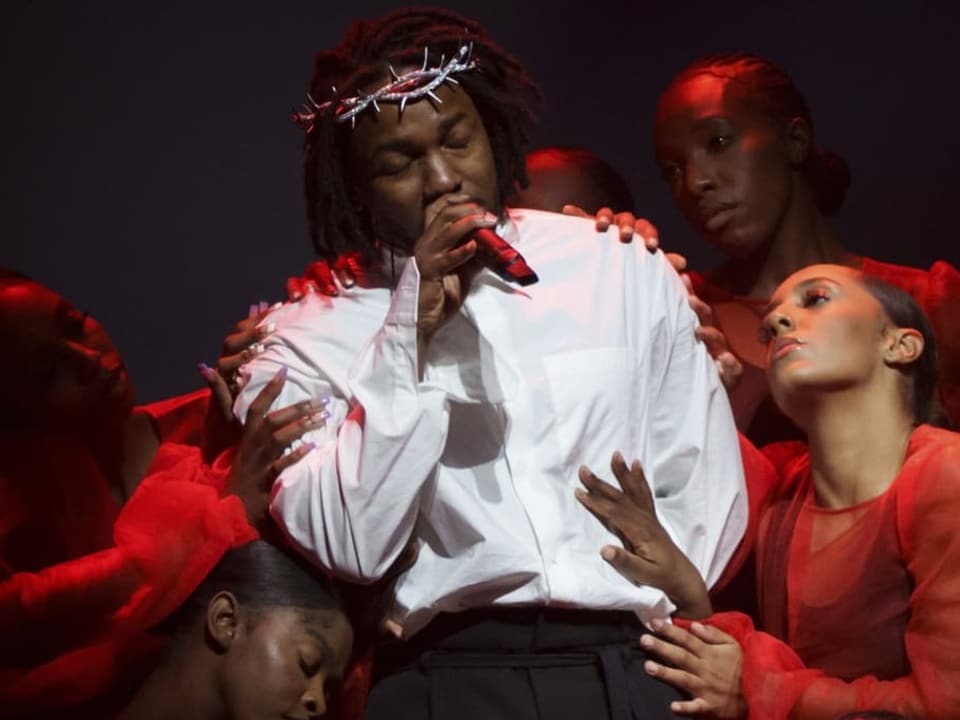 Kendrick Lamar trägt ein weisses Shirt und eine Krone, rund um ihn sind Frauen in rot gekleidet.