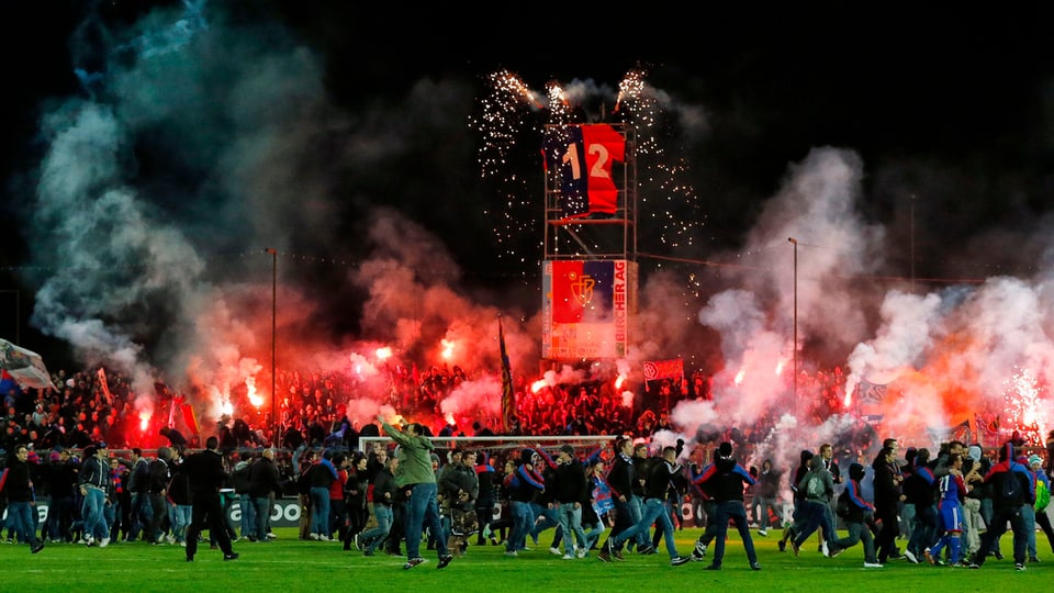 Fans stürmen Spielfeld, im Hintergrund Rauch und rotes Feuer.