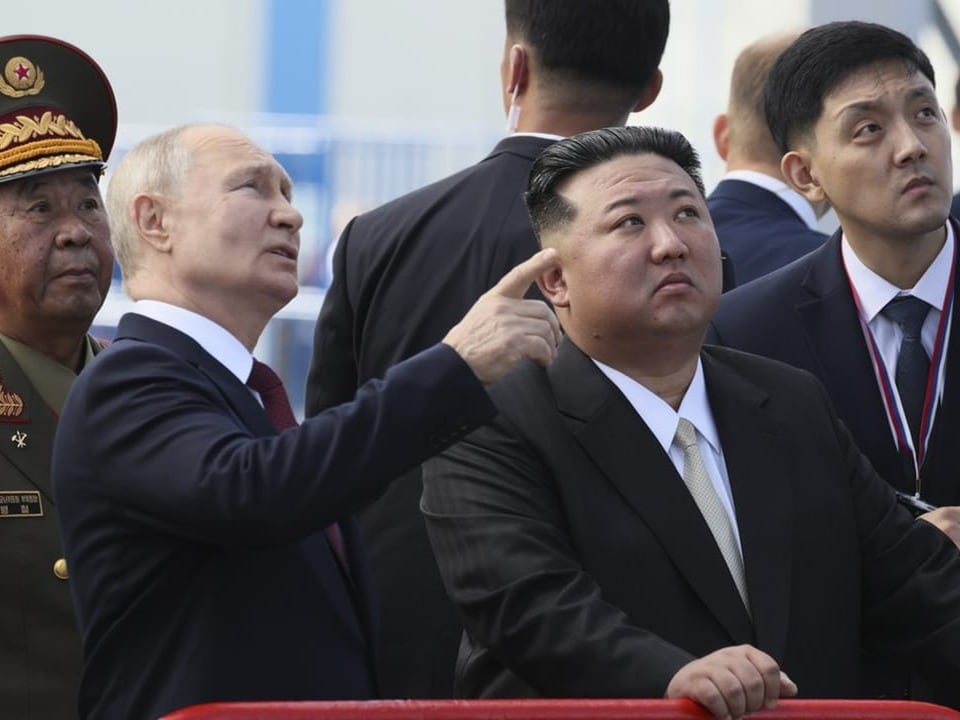 Putin zeigt mit dem Finger in die Luft und Kim Jong-un sowie weitere Männer im Hintergrund folgen seinem Blick.