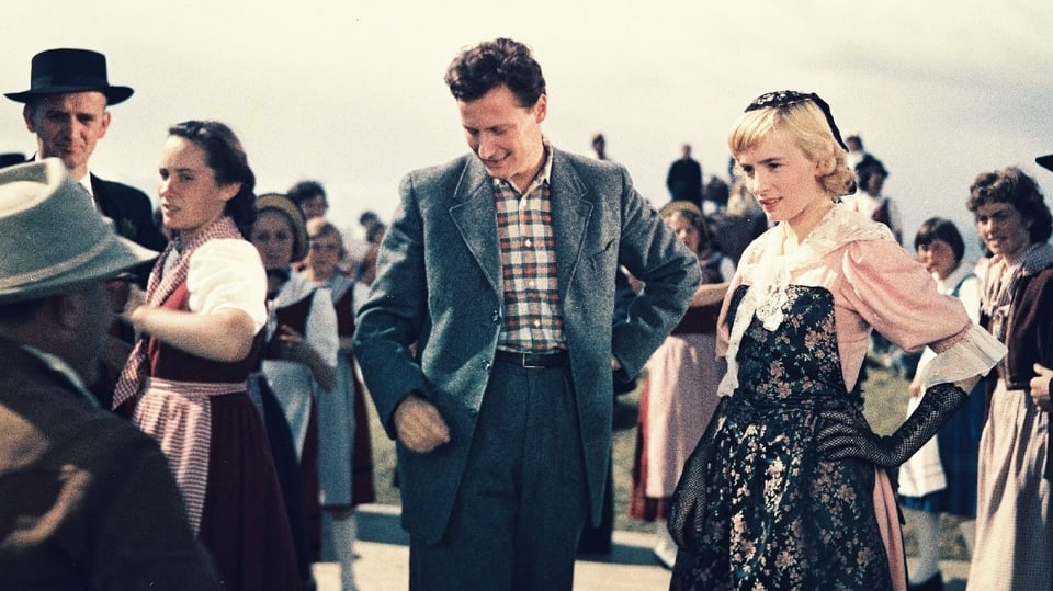 Dominik und Jacqueline an einem Fest. Jacqueline trägt eine Tracht. Um sie herum tanzende Leute.