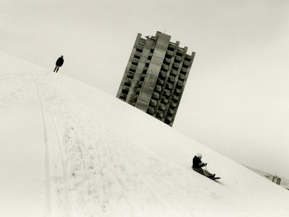 Schwarz-Weiss-Bild: Ein schneebedeckter Hügel mit einem Knaben auf dem Schlitten, am Horizont ein Hochhaus.