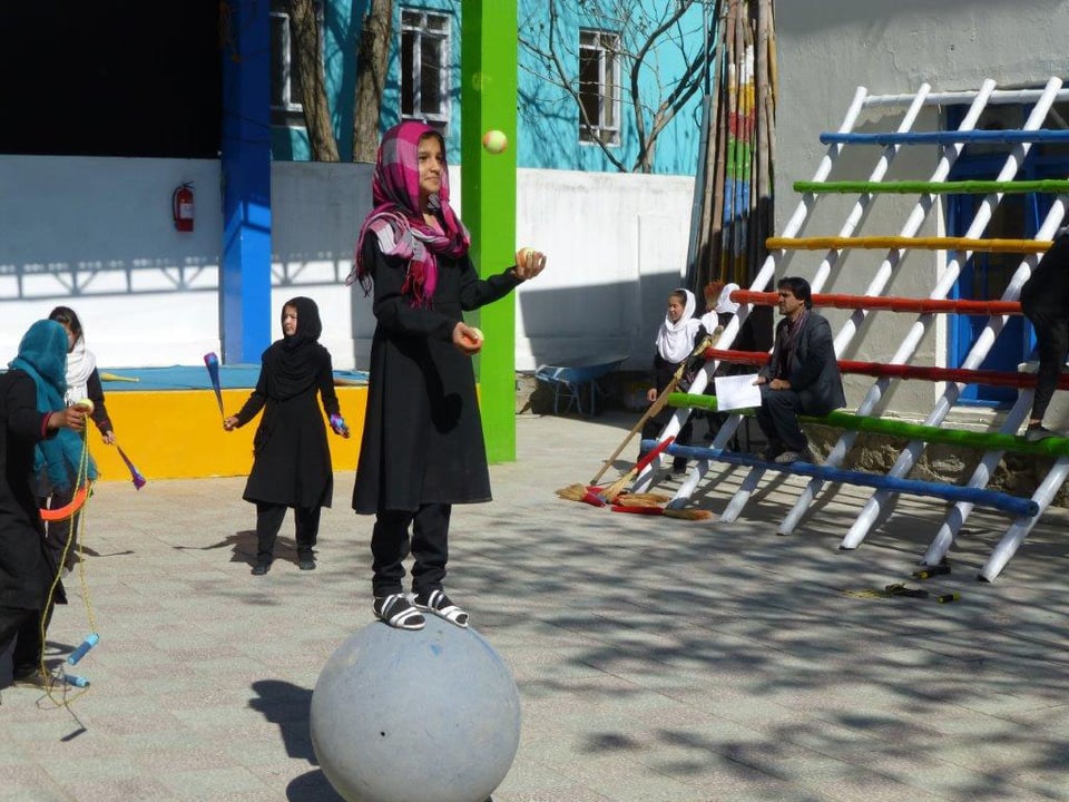 Ein Mädchen steht auf einem Ball und jongliert dazu mit drei Bällen.