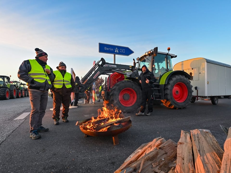 Männer mit Leuchtwesten stehen bei einer Feuerschale, im Hintergrund Traktoren